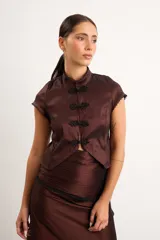 Top de tafeta elastizada con inspiración oriental, color marrón, cuello mao y cierre frontal con nudos chinos y broches. Presenta mangas cortas con detalles de puntilla negra en los bordes.