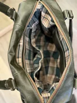 Bolso maternal de cuero sintético color verde oliva con textura rugosa. Presenta múltiples bolsillos exteriores con cierres metálicos y tiradores de cuero color suela, además de bolsillos laterales elásticos. Incluye asas superiores y correa ajustable extraíble.