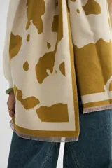 Bufanda rectangular de tejido de punto, con diseño estampado de manchas abstractas en color mostaza sobre fondo crudo y terminación con flecos cortos.