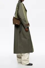 Bolso bandolera de gabardina color marrón, con diseño compacto, cierre de cremallera y asa de tejido ajustable. Presenta el logo de la marca aplicado en la parte frontal.