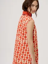 Vestido midi camisero sin mangas, de corte recto y holgado. Cuenta con cuello camisero liso en color naranja y cierre frontal parcial con botones. Presenta un estampado geométrico psicodélico en tonos naranja y beige.
