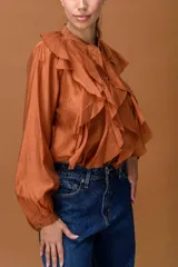 Camisa de color terracota con cuello mao, cierre frontal de botones y volados verticales a lo largo de la pechera. Presenta mangas largas abullonadas con puños ajustados.