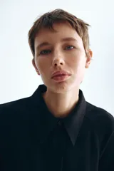 Vestido midi negro con cuello camisero y tapeta con botones ocultos, de mangas largas raglán con puños anchos y costura fruncida en la cadera con falda de vuelo leve.