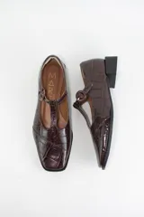 Zapatos estilo Mary Jane de cuero con acabado acharolado y textura efecto croco en color marrón oscuro. Presentan punta cuadrada, cierre con tira en el empeine y taco cuadrado bajo.