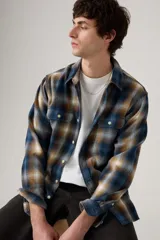 Camisa de franela a cuadros en tonos azul, marrón y blanco, con cuello clásico, cierre de botones frontales y dos bolsillos con solapa en el pecho.