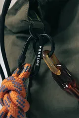 Bolso tipo tote en color verde oliva, confeccionado en tela técnica arrugada. Cuenta con una correa de hombro estilo cuerda en tonos naranja y azul con nudos decorativos, y una cadena decorativa de eslabones de resina en color carey. Presenta cierre superior y detalles de cordones laterales.