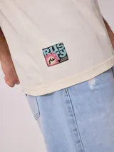 Remera holgada de algodón color beige, de manga corta y cuello redondo, con un gran estampado colorido en la espalda con diseño de olas y la inscripción 'Rusty Tribe'.