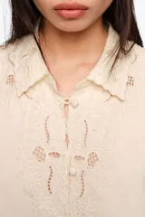 Blusa color beige, de manga corta, con cuello clásico, cierre frontal con botones revestidos y detalles de bordados calados en el frente, cuello, mangas y dobladillo.