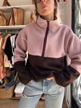 Buzo de polar tipo corderito con diseño de bloques de color en tonos rosa y marrón chocolate. Presenta cuello alto con cierre frontal parcial y un aplique circular en el pecho.