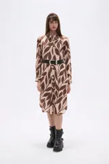 Vestido tipo camisa de corte holgado, con cuello clásico, cierre frontal de botones y mangas largas. Presenta un estampado de hojas en tonos marrones y beige.