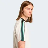 Camiseta Adidas Originals de corte holgado, color blanco con cuello redondo acanalado en negro. Presenta las icónicas tres tiras en los hombros en color negro y verde, y el logo de la marca bordado en el centro del pecho.