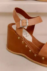 Sandalias de plataforma y taco alto confeccionadas en cuero vacuno marrón, con tira ancha en el empeine, tira ajustable con hebilla en el tobillo y detalles de tachas decorativas en la base.