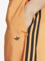 Pantalón deportivo Adidas de corte holgado con diseño degradado en tonos naranja y marrón. Presenta las tres rayas negras características de la marca en los laterales y logo bordado en la parte superior.