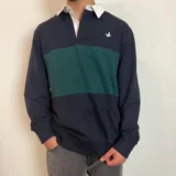 Buzo tipo polo de rugby con cuello blanco, manga larga y puños acanalados. Presenta un diseño de bloques de color en azul marino y verde, con un pequeño logo bordado en el pecho.