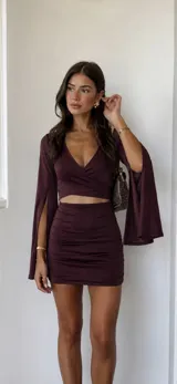 Vestido corto color uva, de estilo dos piezas con efecto cut-out en la cintura. Presenta un top cruzado con escote en V y mangas largas acampanadas con abertura, y una falda ajustada con frunces laterales.