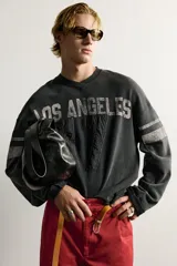 Sudadera cropped fit de color negro con efecto lavado vintage, cuello en V y manga larga. Presenta estampado con texto "LOS ANGELES" y el número "17" en parches acolchados en el frente, además de bandas grises en las mangas.