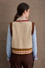 Chaleco de punto color beige con cuello bebé, bordado en rojo, cierre frontal de botones y detalles en marrón y rojo en el borde inferior y bolsillo.