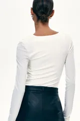 Body de manga larga en punto elástico color crema, con escote drapeado y hombreras. Presenta costuras fruncidas para realzar el efecto drapeado, refuerzo interior con botones a presión y cobertura media en la parte posterior.