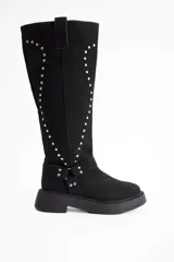 Bota de caña alta confeccionada en cuero vacuno, color negro, con diseño de tachas metálicas dispuestas en forma de arco a lo largo de la caña y correa decorativa con hebilla en la zona del tobillo. Presenta suela track de goma robusta.