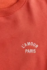 Buzo oversized de cuello redondo y manga larga, en color coral, con el texto 'L'AMOUR PARIS' bordado en el pecho en color blanco.