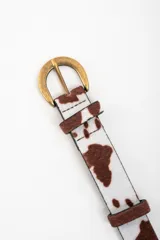 Cinto con estampado de vaca en tonos marrones y blancos, con hebilla metálica dorada envejecida.