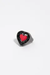 Anillo de metal con diseño en forma de corazón, acabado oscuro y detalle de esmalte rojo en el centro rodeado por un borde negro con letras grabadas.