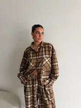 Camisa de viyela con estampado de cuadros en tonos marrones, beige y negro. Presenta cuello clásico, cierre frontal con botones, mangas largas con puños abotonados y dos bolsillos delanteros con solapa.