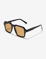 Lentes de sol Hawkers modelo Lash Eco en colaboración con Smiley. Presentan un marco cuadrado de color negro con acabado brillante y cristales en tono ámbar. Incluyen detalles metálicos en las esquinas y el logo de Smiley grabado en las patillas.