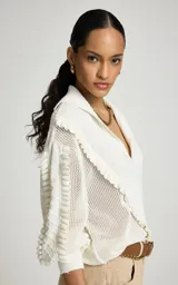 Sweater calado de punto color blanco, de corte recto con amplio cuello tipo polo y escote en V. Presenta mangas largas con volumen, puños ajustados y detalles de volados texturizados en la parte delantera y en las mangas.