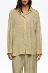 Camisa larga plisada de color verde, confeccionada en tejido ligero. Presenta cuello camisero, cierre frontal con botones, mangas largas con puños abotonados, bolsillo plastrón en el pecho y bajo redondeado, combinando un diseño fluido y versátil.