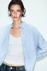 Camisa de manga larga con patrón de rayas verticales en tonos celeste y blanco, cuello con solapa abotonada y un bolsillo aplicado en el pecho. Cierre frontal con botones blancos.