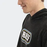 Canguro con capucha de estilo urbano, color negro con efecto lavado. Presenta un estampado gráfico central con el logo de la marca Reef enmarcado en un óvalo con líneas azules y amarillas, y cuenta con un bolsillo frontal tipo canguro.