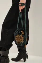 Cartera tipo saco de terciopelo verde oscuro con bordados florales en mostacillas doradas. Cuenta con cierre de cordón ajustable y manija larga trenzada en color verde, con detalles de borlas doradas en los extremos.