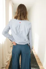 Camisa celeste a rayas verticales blancas, de corte clásico con cuello camisero, mangas largas y cierre frontal con botones rojos. Presenta costuras en contraste y un corazón bordado en la espalda.