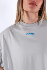 Remera oversize de algodón color gris claro, con cuello redondo y manga corta. Presenta un pequeño logo bordado en azul en el pecho.