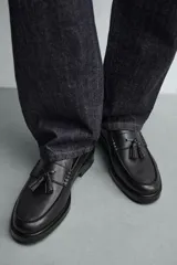 Mocasín de vestir color marrón oscuro, con parte superior lisa, detalle de antifaz con borlas en el empeine y horma redonda. Suela de goma a tono.