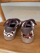 Championes urbanos con diseño de estampado animal print tipo vaca, confeccionados en cuero con textura de pelo. Presentan cordones marrones, suela de goma en tono tostado y detalles en color marrón oscuro en el cuello y lengüeta.