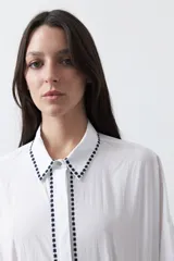 Camisa blanca de manga larga con detalles bordados en contraste a lo largo de la botonadura, el cuello y los puños. Presenta una silueta relajada y un diseño clásico con un toque artesanal.