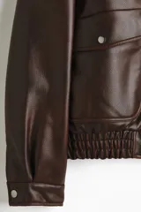 Campera de estilo aviador confeccionada en tejido revestido con acabado de cuero sintético. Presenta cuello de sherpa en contraste, cierre frontal de cremallera, hombros caídos y bolsillos delanteros de parche con solapa y botón a presión. Incluye elástico fruncido en el bajo y puños ajustables con botón a presión.