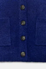 Chaqueta de punto de corte cropped y manga larga, con cuello redondo y cierre frontal de botones. Presenta dos bolsillos de plastrón en la parte delantera y acabados en rib.
