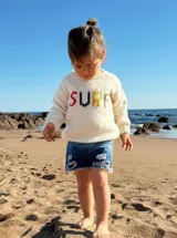 Sweater tejido a mano en lana merino color crudo con cuello redondo y mangas largas. Presenta la palabra "SURF" bordada en el frente con letras de colores.