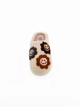 Pantuflas de estar por casa tipo mule, confeccionadas en tejido de peluche color crema. Presentan un diseño con apliques bordados de caritas sonrientes en tonos marrones, bordó y lila. Cuentan con una suela plana de goma y forro interior suave.
