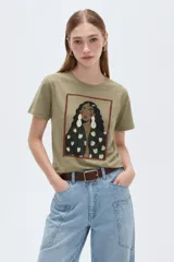 Remera de algodón color verde militar, de manga corta, con cuello redondo y diseño ajustado. Cuenta con un estampado frontal central enmarcado que ilustra a una mujer con grandes aretes.