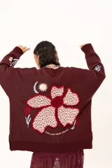 Saco de punto color vino con diseño holgado, cuello redondo y mangas largas. Presenta un gran bordado central en la espalda con forma de flor en tonos crema y rojo, acompañado de un pequeño bordado de sol y una frase bordada en la parte inferior.
