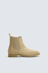 Bota estilo Chelsea de piel con acabado serraje color beige, con elásticos laterales para facilitar el calce, horma redonda y suela a tono con ligero tacón.
