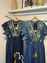 Vestido largo de denim azul con escote en V, mangas cortas con volados y cordón ajustable con borlas en la cintura. Presenta bordados detallados en el pecho y apliques grandes de flores con flecos en la parte inferior de la falda.