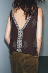 Top sin mangas color negro con escote pico profundo. Presenta bordados florales y un panel central con diseño geométrico, ambos realizados con aplicación de abalorios plateados o blancos.