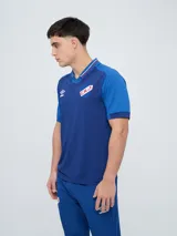 Remera polo deportiva Umbro del Club Nacional de Football, color azul con cuello en V y detalles en rojo y blanco. Incluye el logo de Umbro y el escudo del club.