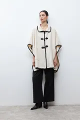 Capa color crudo con cuello camisero y ribetes negros. Cierre frontal con alamares negros y bolsillos laterales.