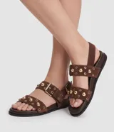 Sandalias planas Satinato de estilo sport sandal, confeccionadas en material sintético efecto gamuza color marrón. Presentan dos tiras cruzadas en el empeine decoradas con tachuelas semiesféricas doradas y ojales metálicos, además de una tira trasera con hebilla ajustable.